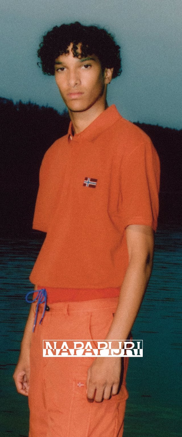 Camisa polo naranja con un pequeño parche de bandera, combinada con pantalones naranja a juego y acentos de cordón azul. El fondo muestra agua.