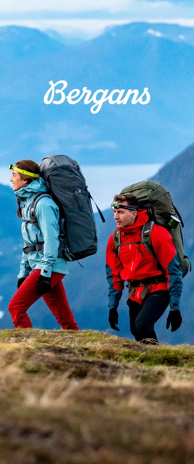 Dos personas hacen senderismo en una pendiente cubierta de hierba, vistiendo ropa de exterior en capas: una chaqueta azul y pantalones rojos, una chaqueta roja y mochilas verdes.