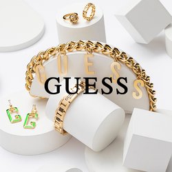 Zlaté šperky Guess, včetně řetízkového náramku, dvou prstenů a zelených náušnic, vystavené na bílých geometrických tvarech na bílém pozadí.