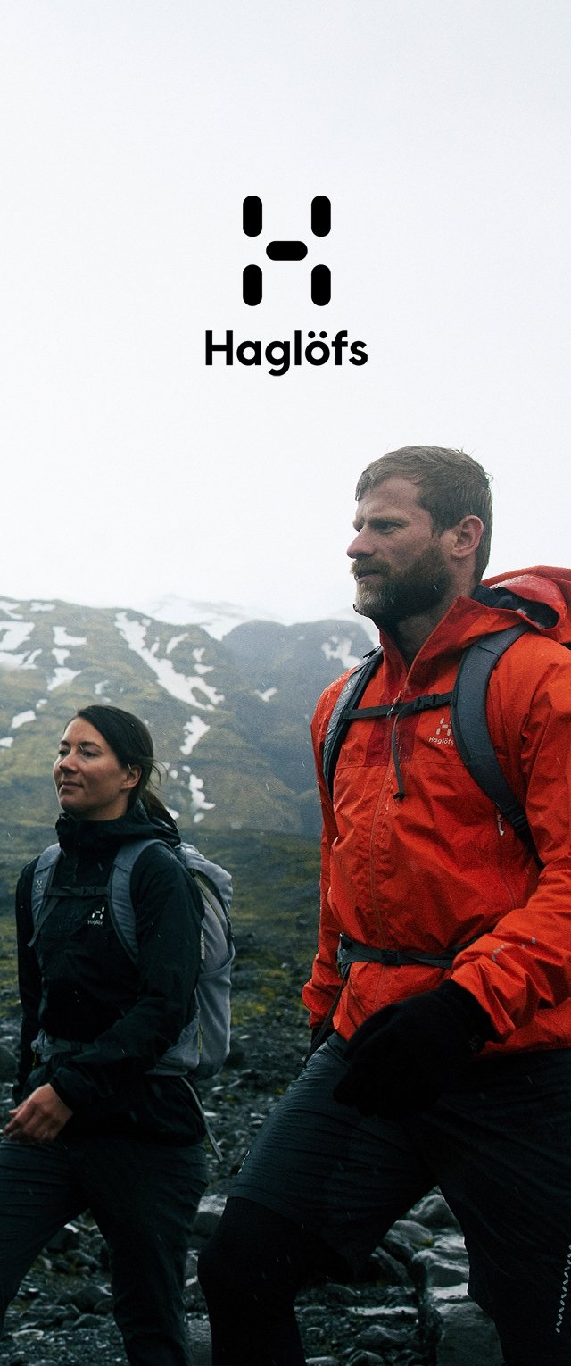 Zwei Wanderer gehen über felsiges Gelände. Einer trägt eine leuchtend orangefarbene Jacke, der andere eine dunkle Jacke. Beide haben Rucksäcke. Im Hintergrund sind verschneite Berge zu sehen.