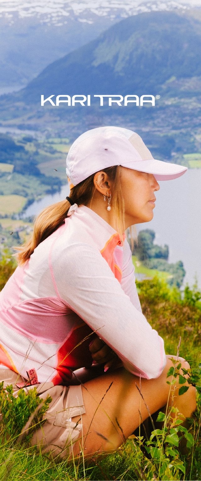 Light pink long-sleeve top with red accents, paired with beige shorts. The person wears a white cap and silver earrings, seated in greenery.