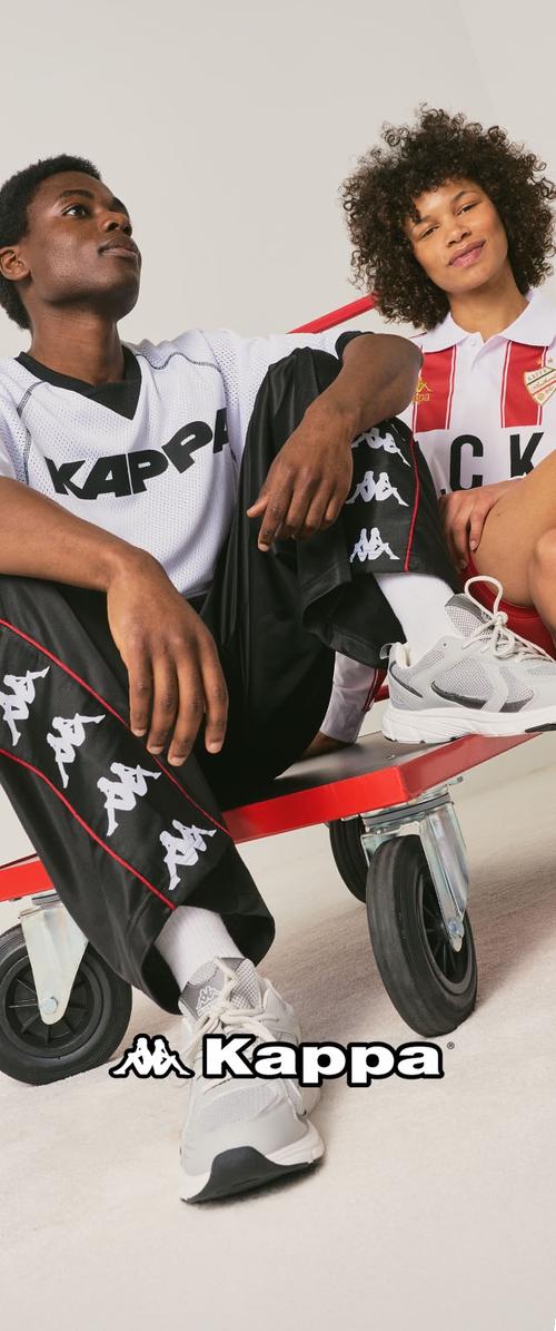 Twee nonchalant zittende modellen die Kappa sportkleding dragen, inclusief logo-bedrukte shirts, broeken en sneakers, op een rood platform op wielen.