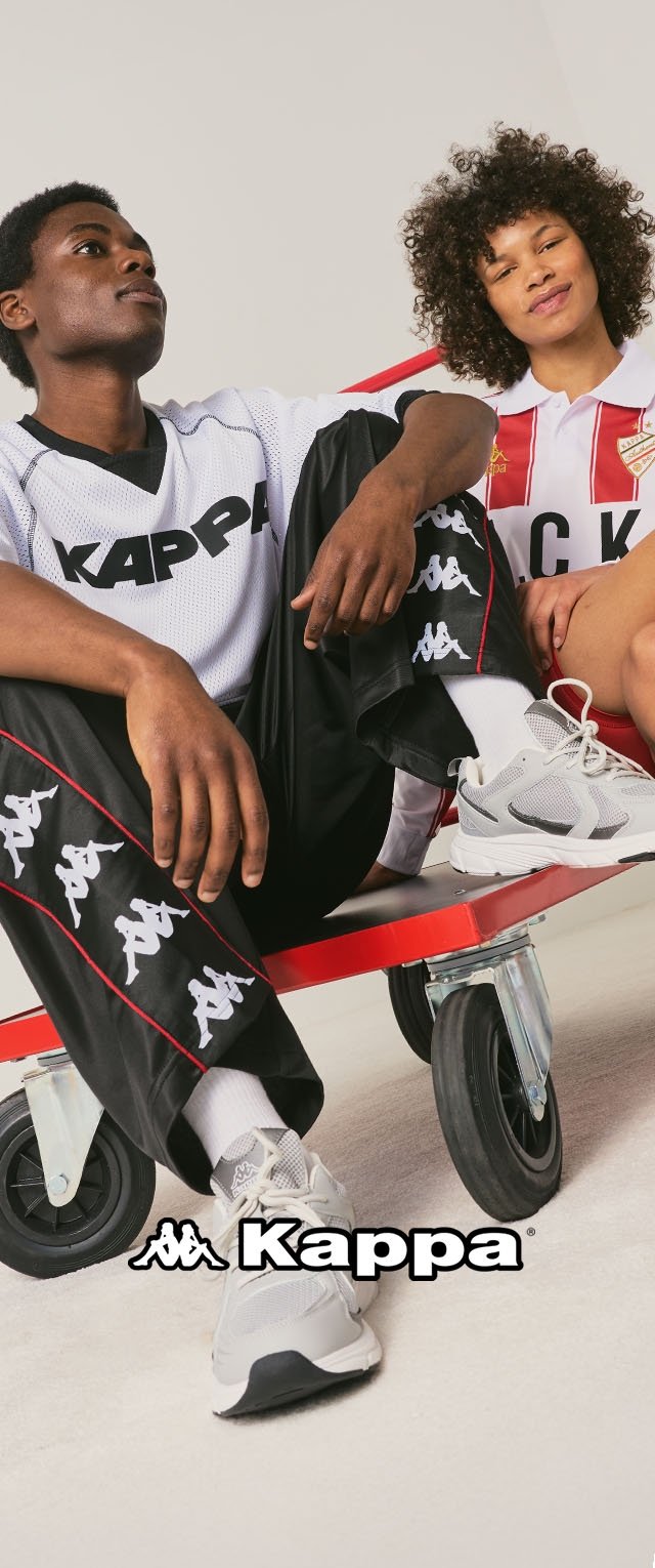 Deux mannequins assis de manière décontractée portant des vêtements de sport Kappa, comprenant des chemises, pantalons et baskets avec logo, sur une plateforme rouge à roulettes.