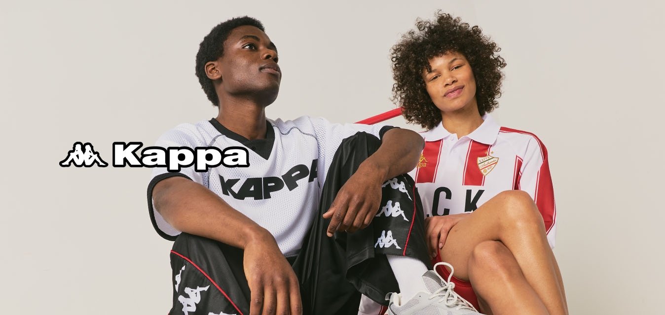 Dva lidé oblečení v sportovním oblečení značky Kappa sedí na červeném plošinovém vozíku před jednobarevným pozadím.