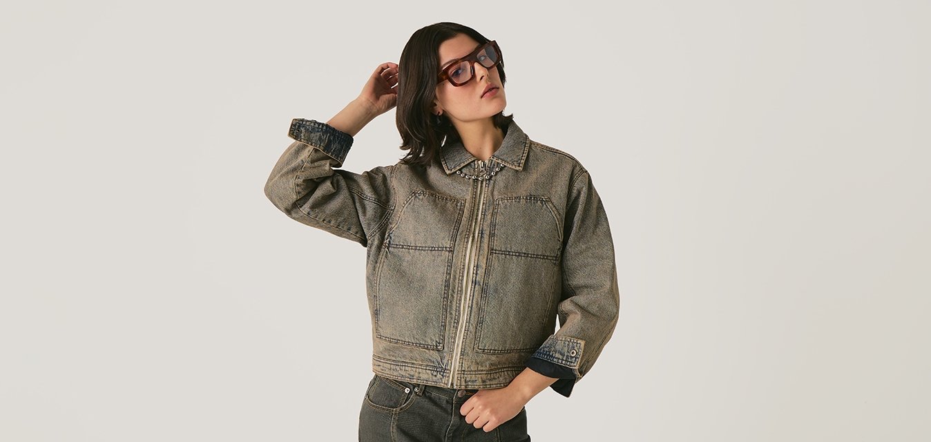 Žena v oversize denimové bundě a odpovídající sukni, upravující si vlasy na neutrálním pozadí.