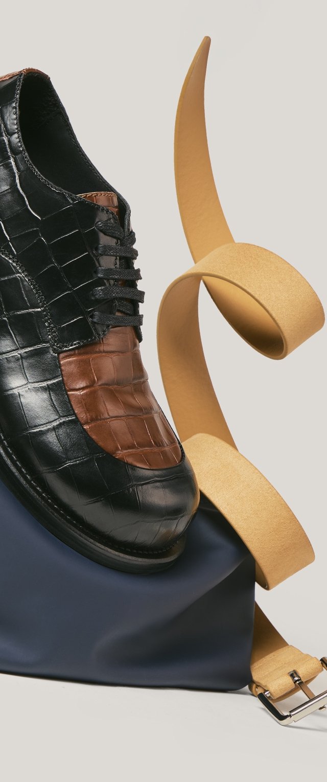 Zapato de cuero negro y marrón con textura de cocodrilo y diseño de cordones, descansando sobre una bolsa azul marino con una correa enrollada amarilla.