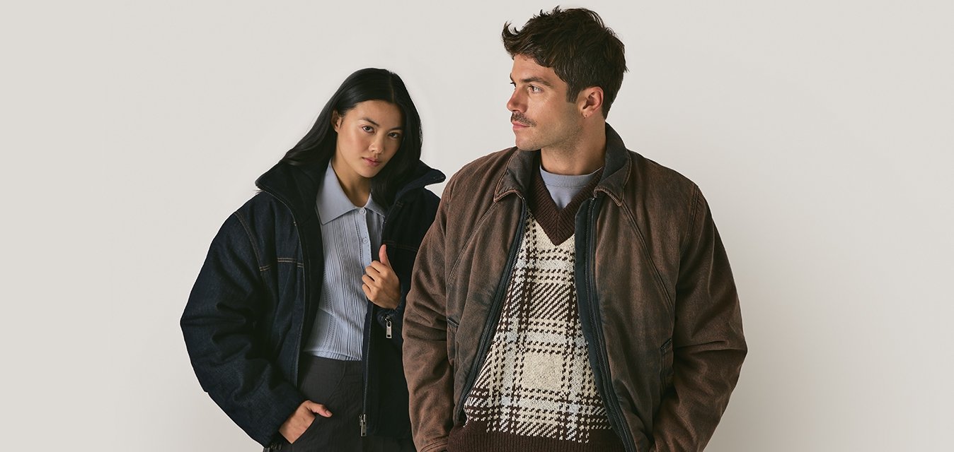 Dva jednotlivci mají na sobě bundy: jeden v tmavé denimové péřové bundě a druhý v hnědé kožené bundě přes vzorovaný svetr. Oba mají slim fit.