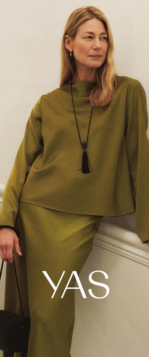 Vrouw met lang blond haar, gekleed in een olijfgroen top met lange mouwen en een rok, met een zwarte ketting met franjes, die een zwarte handtas vasthoudt en leunt op een reling.