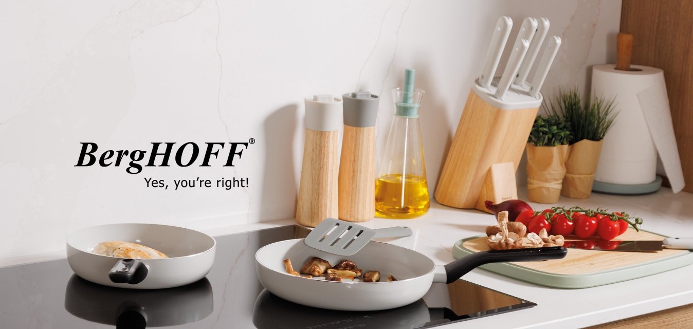 Dvě pánve na černém indukčním sporáku, skříňky ze světlého dřeva, bílá mramorová zástěna a kuchyňské náčiní včetně nože, lahví a rajčat.