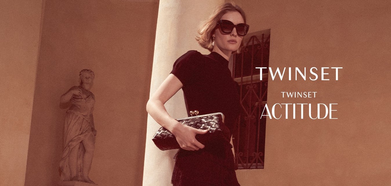 Žena v černých šatech a slunečních brýlích drží psaníčko a kráčí po kamenných schodech vedle sloupu a klasické sochy.