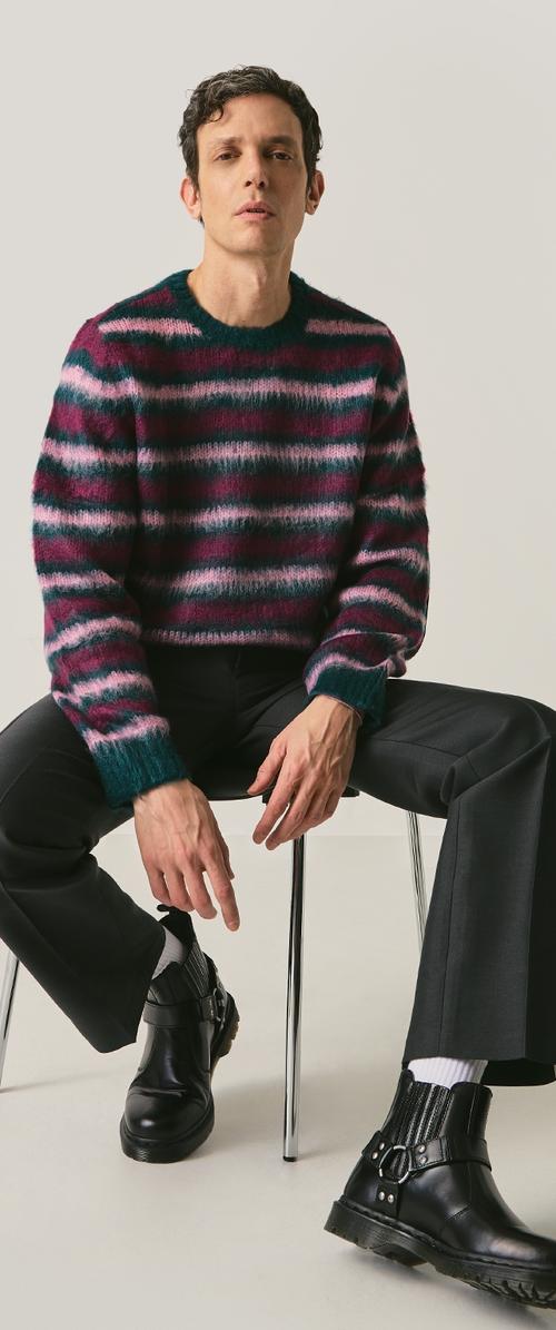 Pulover tricotat cu dungi în nuanțe de burgundy, teal și roz; gât rotund și mâneci lungi, asortat cu pantaloni negri și cizme negre masive.