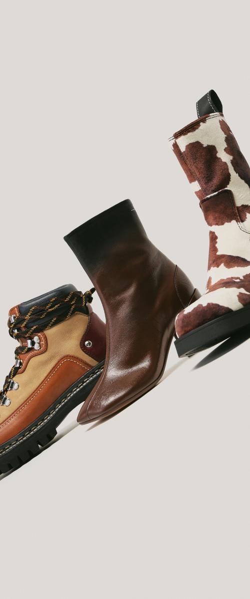Tres pares de botas: el de la izquierda es de cuero marrón claro y oscuro con cordones negros, el del medio es de cuero marrón brillante, y el de la derecha es blanco con estampado de vaca marrón.