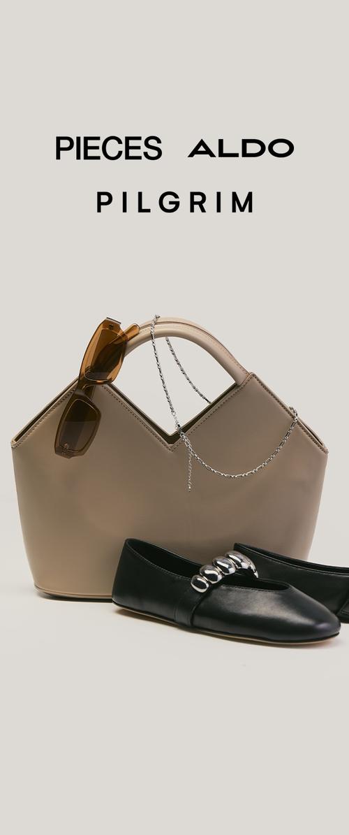 Beige handtas met ketting en bruine zonnebril, gecombineerd met zwarte platte schoenen met zilveren ronde versieringen op een beige achtergrond.