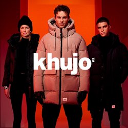 Tři lidé v zimních kabátech a botách stojí v červeně osvětleném koridoru s geometrickými stěnami a podlahou.