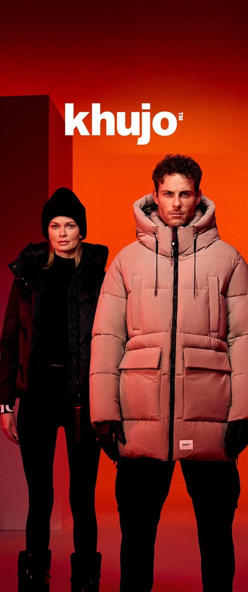 Mann in beigefarbener Steppjacke und Frau in schwarzem Wintermantel mit Mütze stehen vor einem orangefarbenen Hintergrund mit dem "khujo"-Logo darüber.