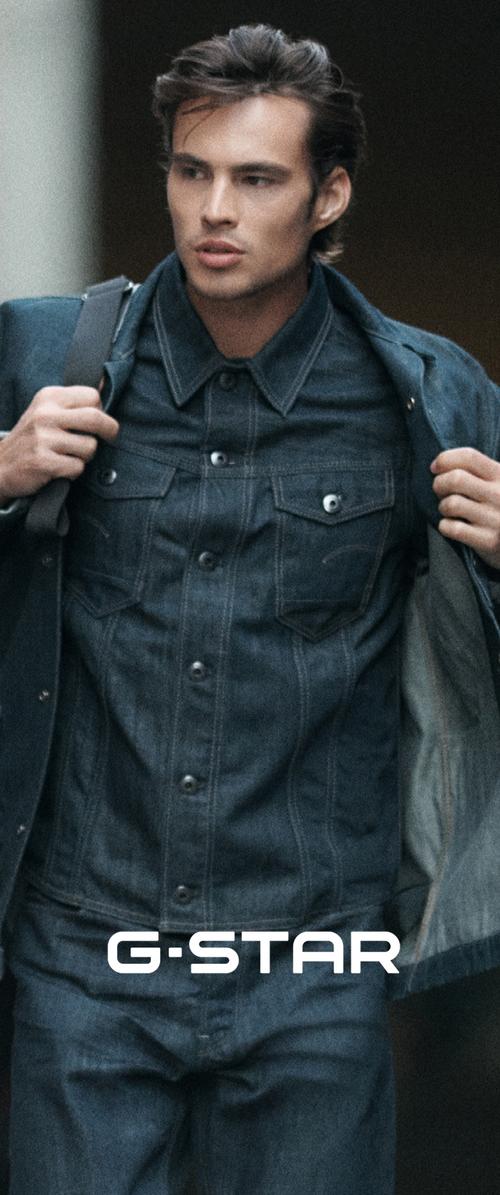 Joven ajustándose una chaqueta de denim oscuro llevada sobre una camisa y vaqueros de denim a juego, con una correa de mochila sobre el hombro, con el logo de G-STAR.