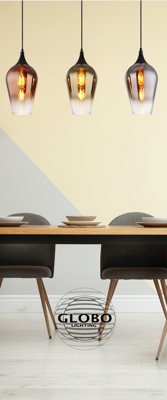 Tres luces colgantes con pantallas de vidrio cilíndricas en ámbar, negro y transparente, colgando sobre una mesa de madera con platos de cerámica.