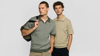 Zwei Männer stehen vor einem schlichten Hintergrund, einer trägt ein grünes Strickpolo mit einer Jacke über der Schulter, der andere ein beiges Strickpolo mit schwarzer Hose.