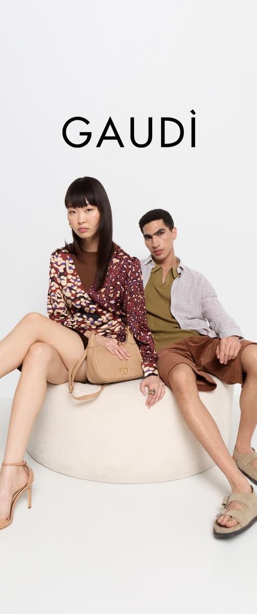 Zwei Models posieren auf einer hellen runden Plattform. Die Frau trägt eine gemusterte Bluse und hält eine beigefarbene Handtasche. Der Mann ist in einem grünen Oberteil und braunen Shorts gekleidet.