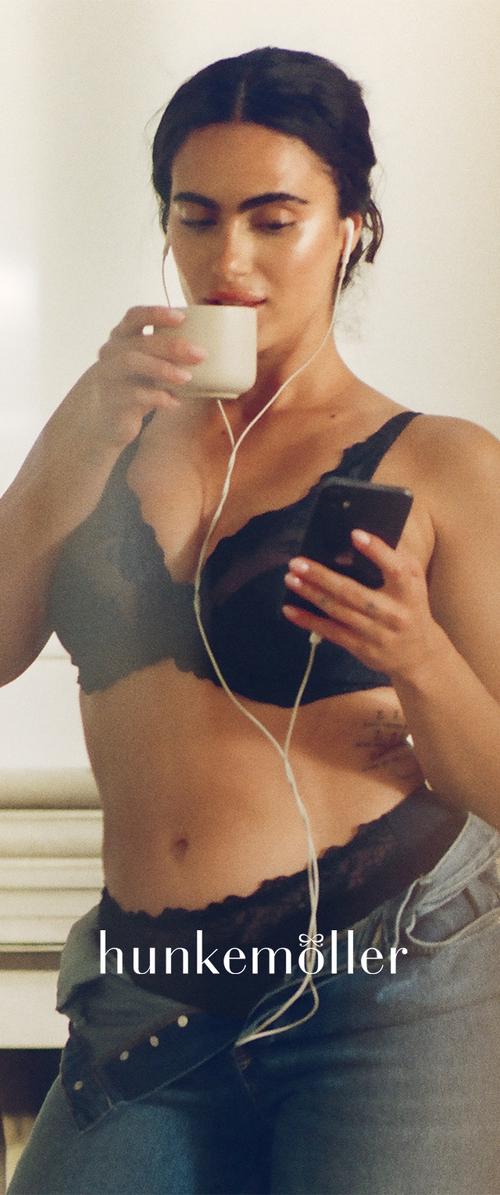Musta pitsinen bralette, jossa on aaltoilevat reunat, yhdistetty farkkuihin. Henkilö pitää valkoista kuppia ja älypuhelinta kädessään, ja hänellä on kuulokkeet korvissa.