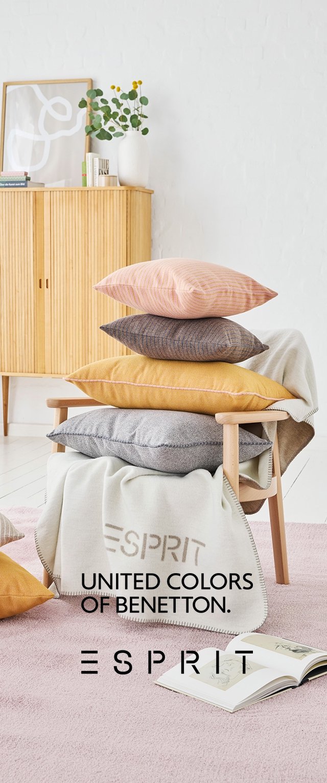 Stapeltje van vijf decoratieve kussens in verschillende kleuren (roze, oranje, grijs, bruin, geel) op een stoel, bedekt met een lichte deken.