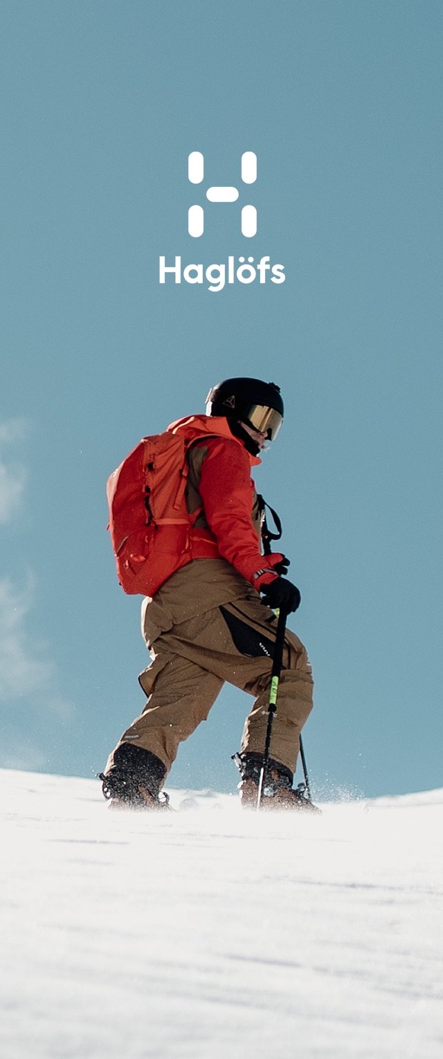 Skifahrer in roter Jacke und beigen Hosen, der einen Skistock hält und mit Rucksack und schwarzem Helm bei blauem Himmel im Schnee geht.