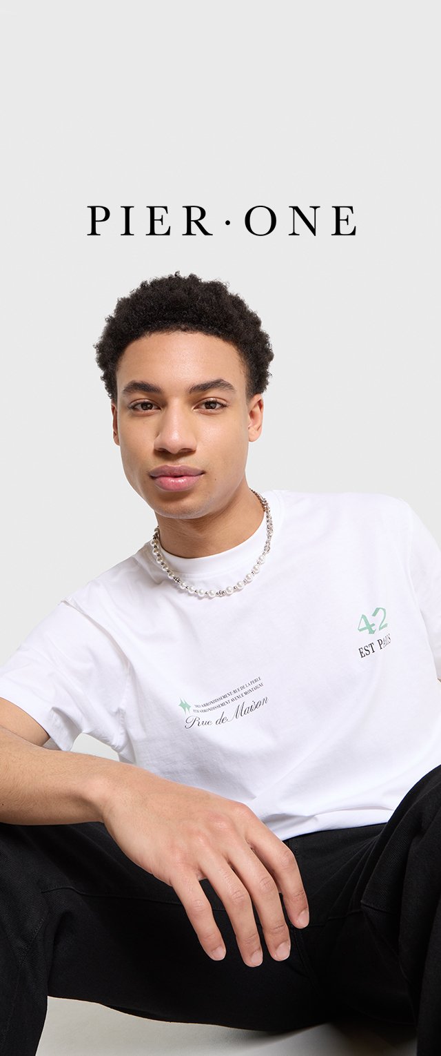 T-shirt bianco con testo e logo verdi, indossato con una collana di perline argentate. Il modello ha i capelli corti e ricci ed è seduto in una postura informale.