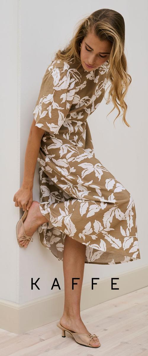 Frau, die einen beigefarbenen Mules-Schuh anpasst, während sie ein hellbraunes Kleid mit weißem Blumenmuster trägt und vor einer weißen Wand auf einem hellen Holzboden steht.