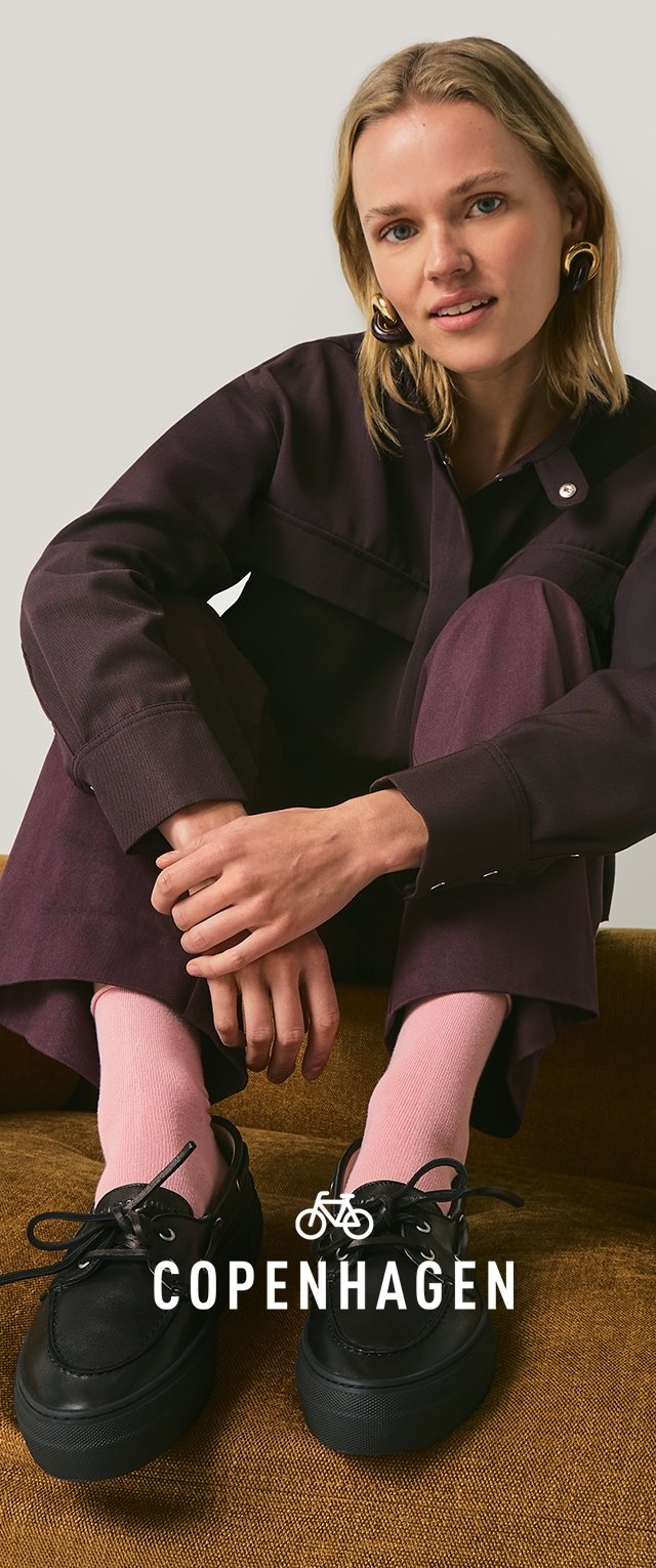 Czarne buty na sznurowadła, jasnoróżowe skarpetki i ciemny strój z krótką kurtką i dopasowanymi spodniami. Siedząca poza na brązowej powierzchni.