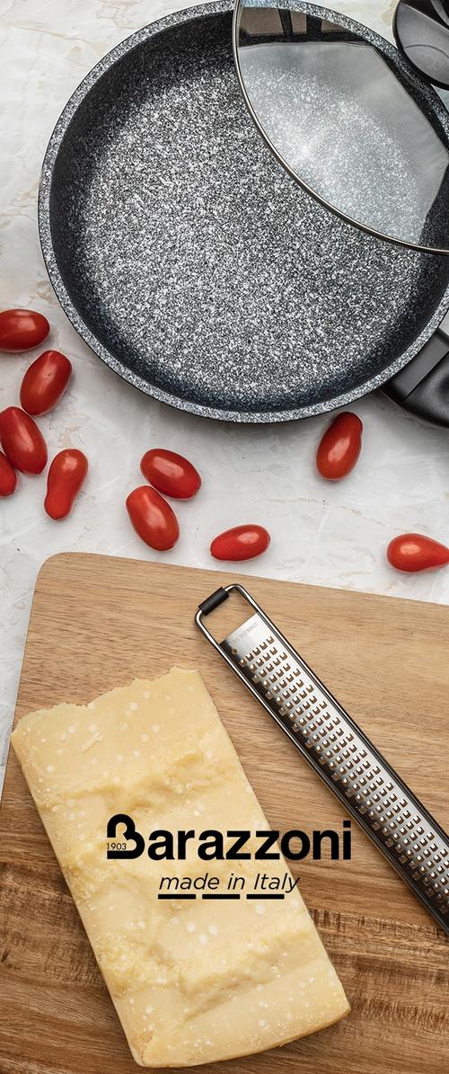 Sartén negra moteada con una tapa de vidrio, rodeada de tomates cherry y un bloque de queso en una tabla de cortar de madera con un rallador.