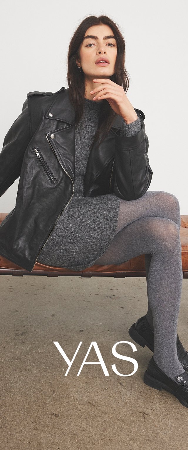 Veste en cuir noir de style motard sur une robe en tricot gris, assortie de collants gris et de mocassins noirs. Modèle assis, mettant en valeur les détails de la tenue.