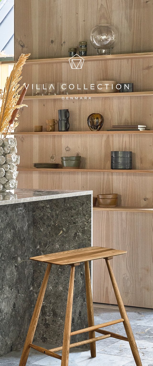 Estantes de madera exhiben varios objetos de vidrio y cerámica sobre una encimera de piedra oscura con un acabado mate. Un taburete de madera se encuentra cerca.