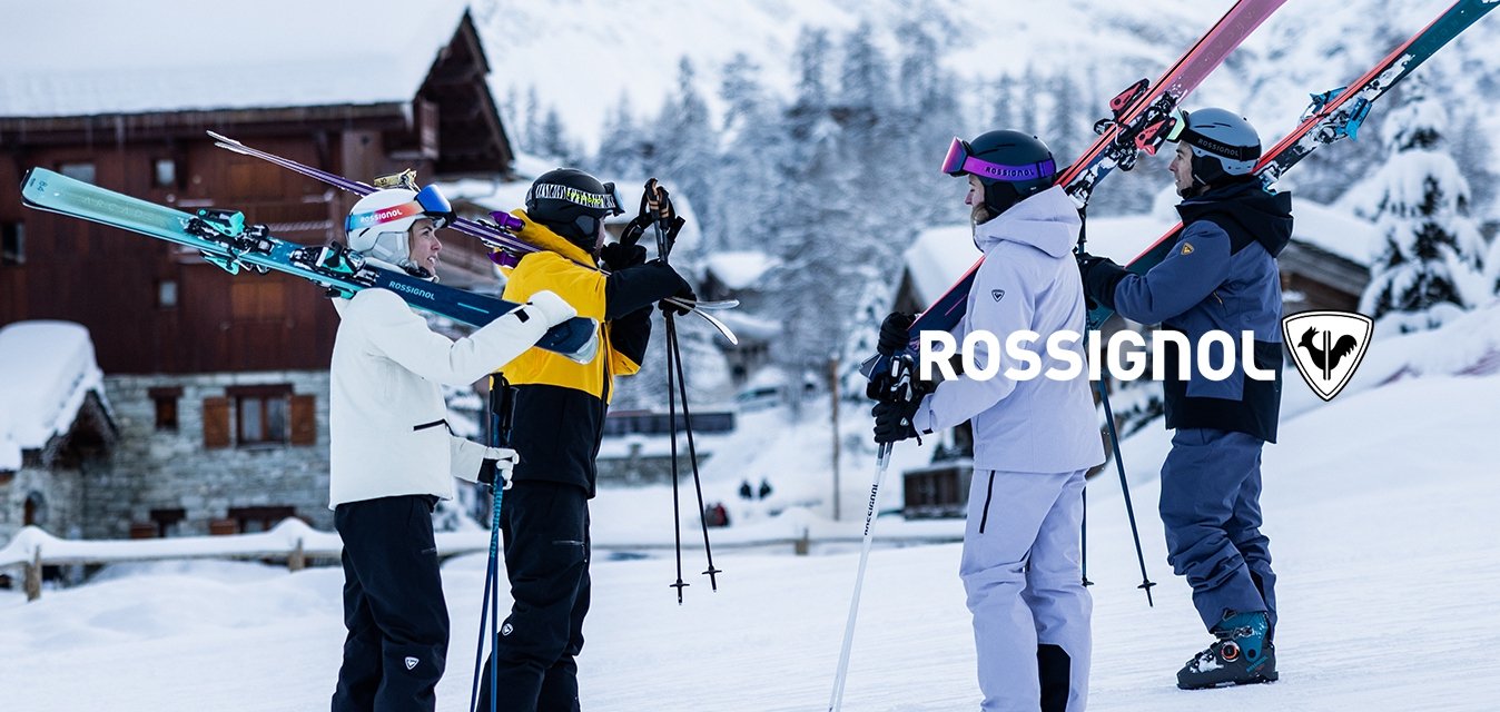 Čtyři lyžaři v zimním oblečení nesou lyže přes ramena, stojí na sněhu poblíž dřevěných chat s zasněženými horami v pozadí.