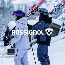 Čtyři lyžaři v zimním oblečení nesou lyže přes ramena, stojí na sněhu poblíž dřevěných chat s zasněženými horami v pozadí.