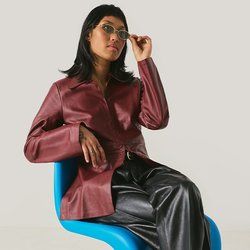 Žena v vínové bundě a černých kalhotách sedí na moderní modré židli a upravuje si sluneční brýle na světlém jednobarevném pozadí.
