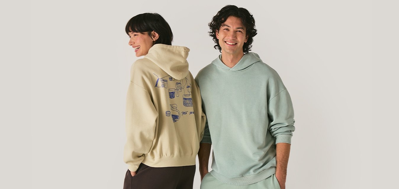 Usmívající se muž a žena oblékající pastelové mikiny a volné kalhoty, stojící vedle sebe na jednobarevném světlém pozadí.