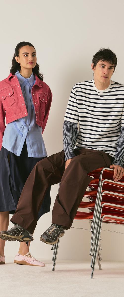 Jeune femme debout vêtue de vêtements décontractés en couches, homme assis sur des chaises rouges empilées, portant une chemise à manches longues rayée et un pantalon marron.
