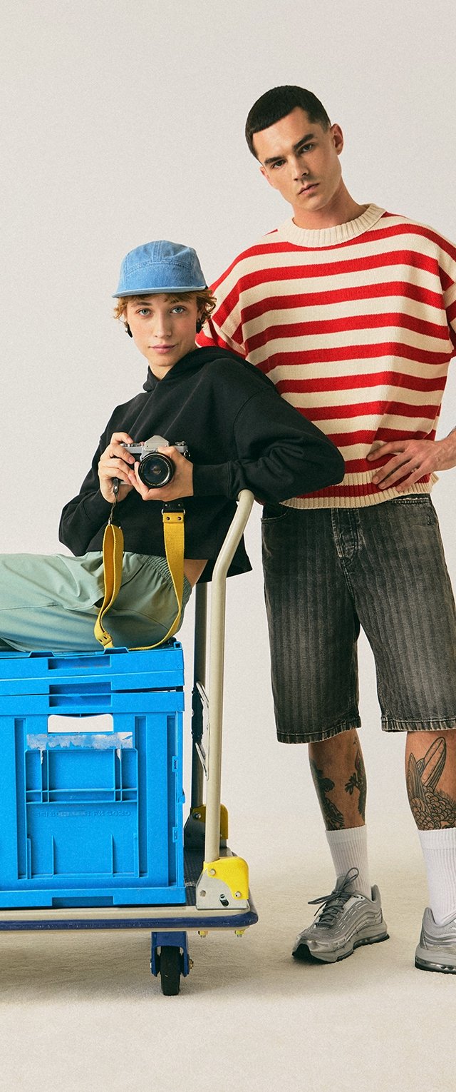 Mladý člověk v modré kšiltovce, který drží fotoaparát, sedí na modré bedně na vozíku, zatímco další stojí vedle v červeném a bílém pruhovaném svetru a šortkách.