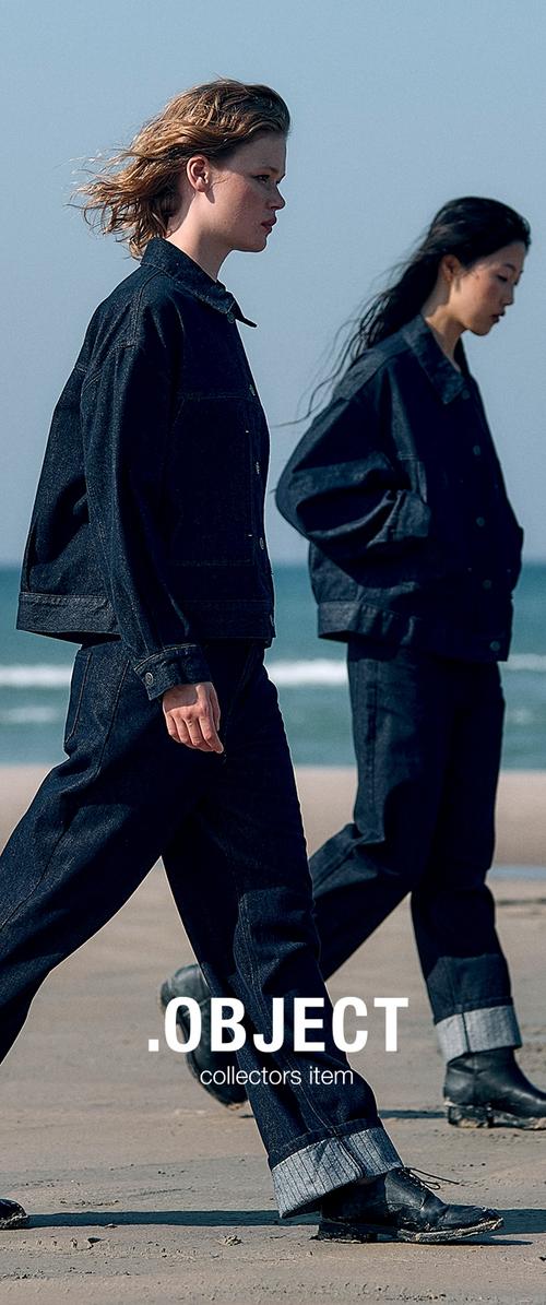 Due modelli camminano su una spiaggia, indossando giacche e pantaloni in denim scuro con risvolti. Le giacche presentano tasche frontali e una vestibilità comoda.