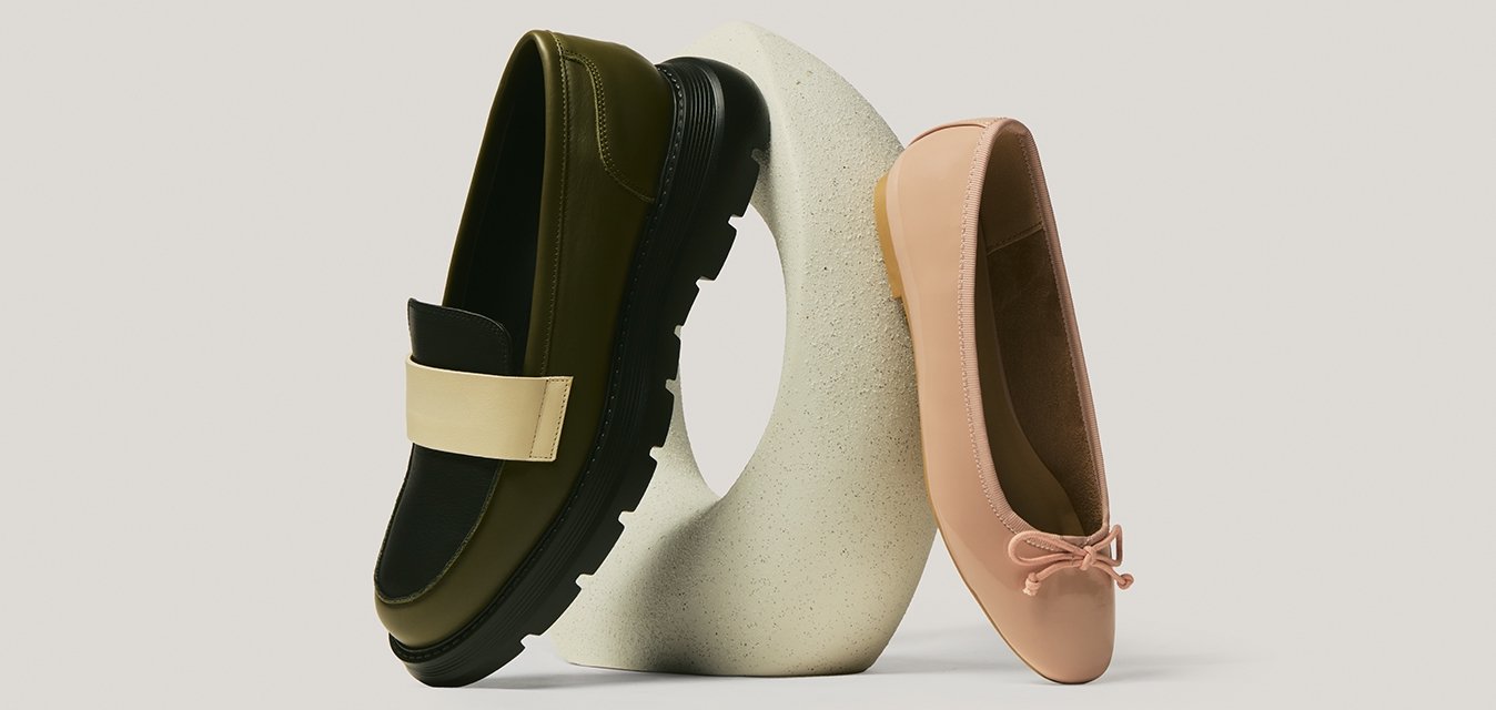 Jedna olivově zelená mokasína s béžovým páskem a jedna světle růžová balerína s mašlí vystavené na zakřiveném bílém kameni.