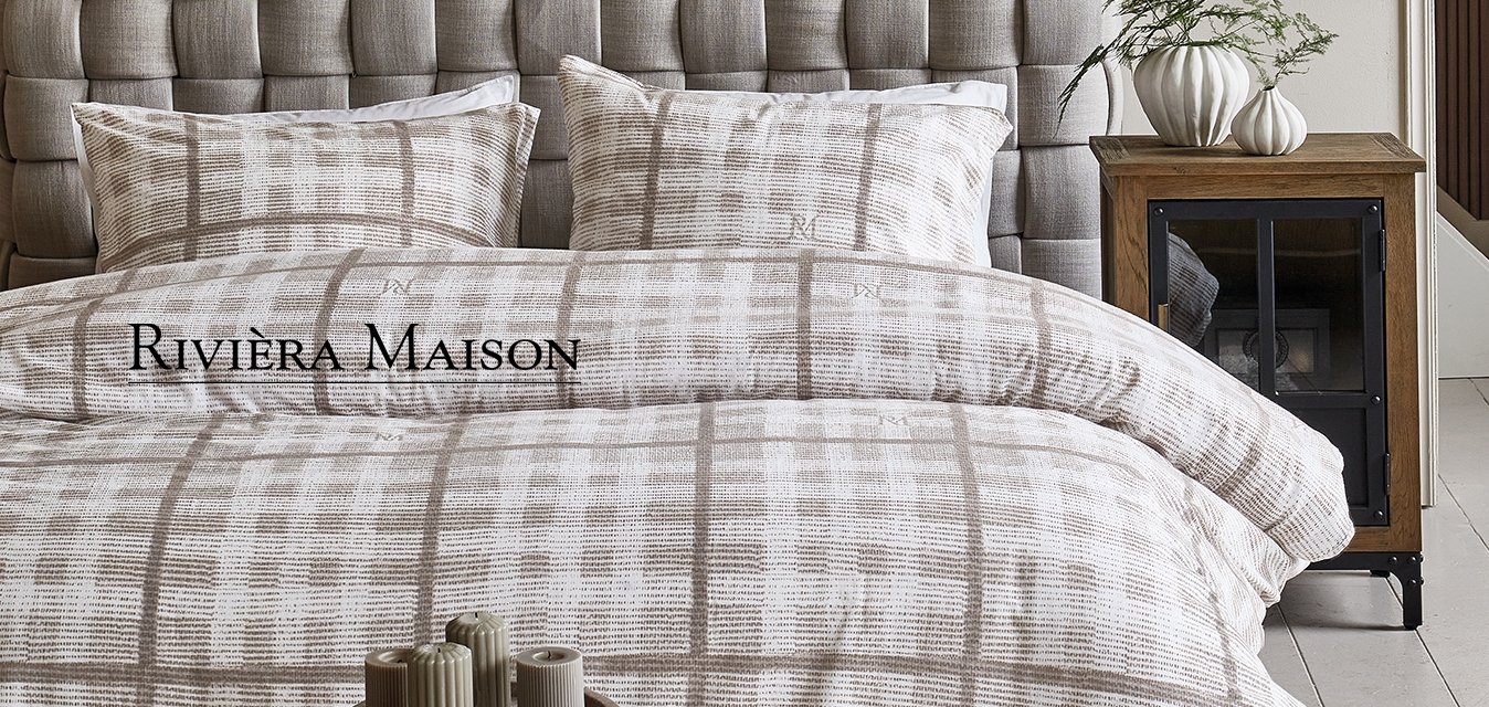 Šedá čalouněná čelní část postele s prošívaným designem, doplněná béžovým a bílým kostkovaným povlečením a vzorovanými polštáři na dřevěném nočním stolku.