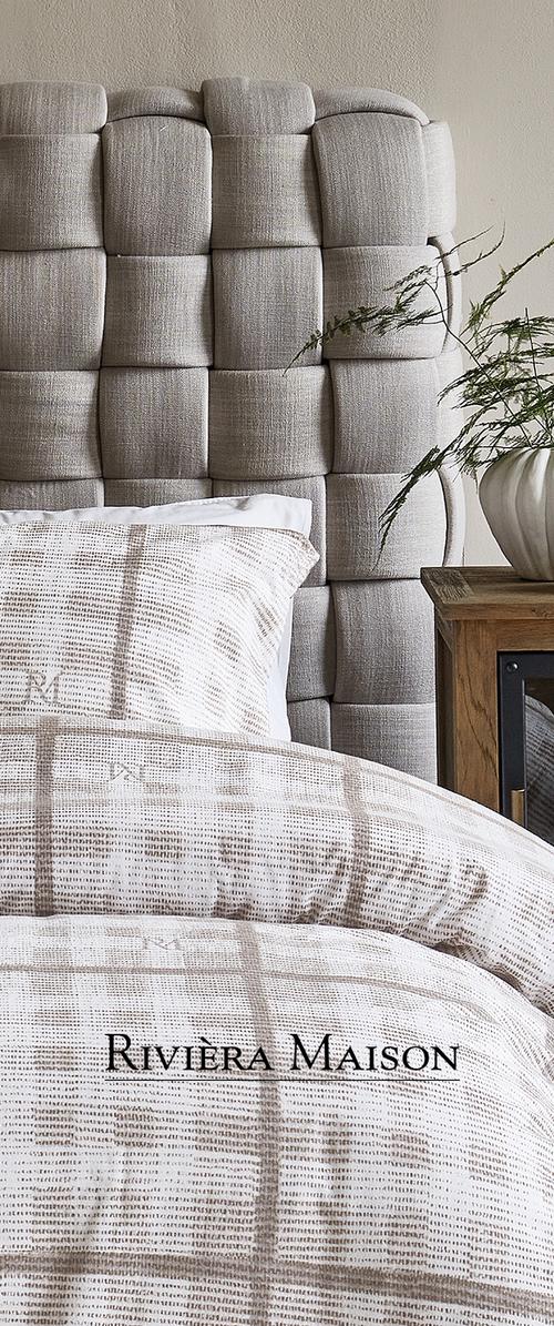 Testata letto beige imbottita con un motivo a griglia, accompagnata da biancheria da letto color crema con un sottile motivo a quadretti. Comodino in legno con una pianta.