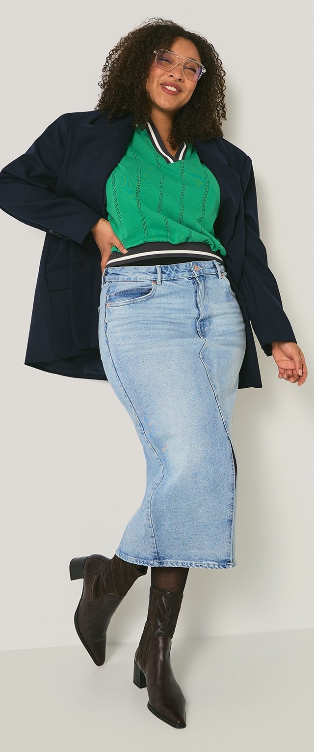 Kobieta o kręconych włosach, nosząca okulary, zielony sweter, granatową marynarkę, jasnoniebieską spódnicę midi z denimu, czarne rajstopy i czarne botki na obcasie, uśmiechająca się.