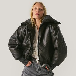 Žena v černé lesklé volné bundě, bílém tričku a širokých pruhovaných kalhotách stojící s rukama v kapsách na jednobarevném pozadí.