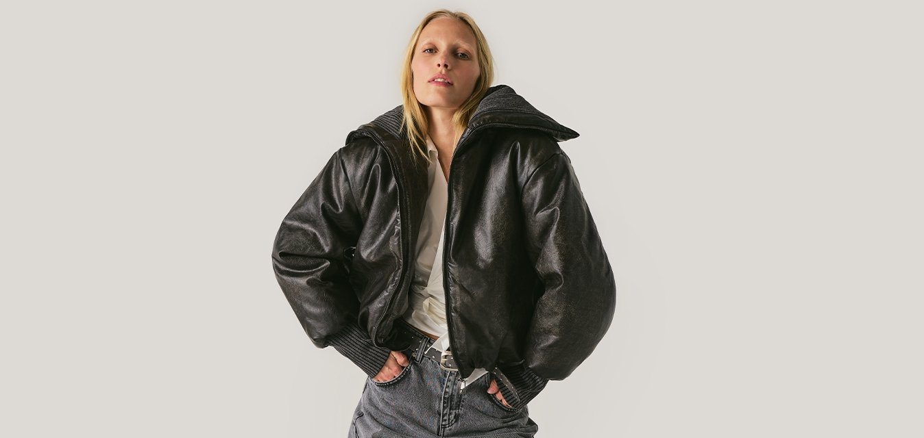 Žena v černé lesklé volné bundě, bílém tričku a širokých pruhovaných kalhotách stojící s rukama v kapsách na jednobarevném pozadí.