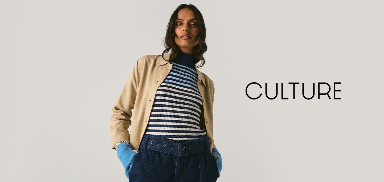 Osoba oblečená v béžové bundě, pruhovaném svetru, širokém pásku, tmavých kalhotách a jasně modrých rukavicích, stojící na neutrálním světlém pozadí.