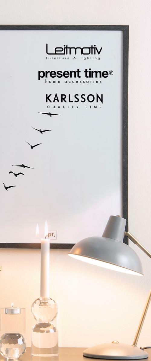 Weißer Wand mit einem schwarz gerahmten Druck von Vögeln im Flug. Im Vordergrund steht ein klarer Kerzenständer und eine graue Schreibtischlampe.