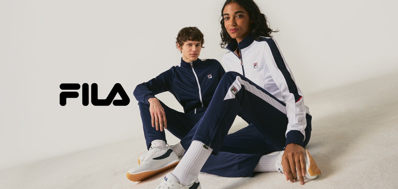 Dvě osoby oblečené ve tmavě modrých a bílých teplákových soupravách značky FILA a bílých teniskách sedí na zemi před jednobarevným světlým pozadím.