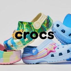 Pět barevných bot Crocs uspořádaných do hravé rovnováhy, přičemž dvě jsou uvnitř nakloněného modrého průhledného kbelíku na bílém pozadí.