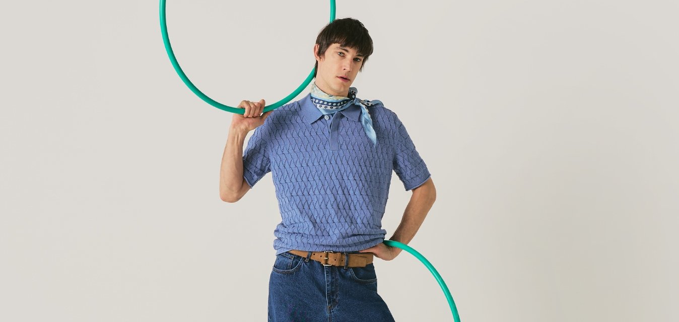 Osoba v modrém svetru a džínách pózuje s zeleným hula hoop, stojící na jednobarevném světlém pozadí.